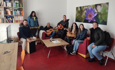 Eine Personengruppe sitzt in einer gemütlichen Sitzecke. Einige Personen halten Musikinstrumente in der Hand.