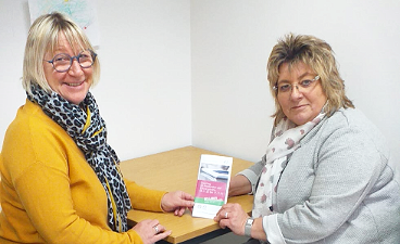 News- Foto: Zwei Frauen posieren an einem Tisch sitzend mit einem Flyer in der Hand